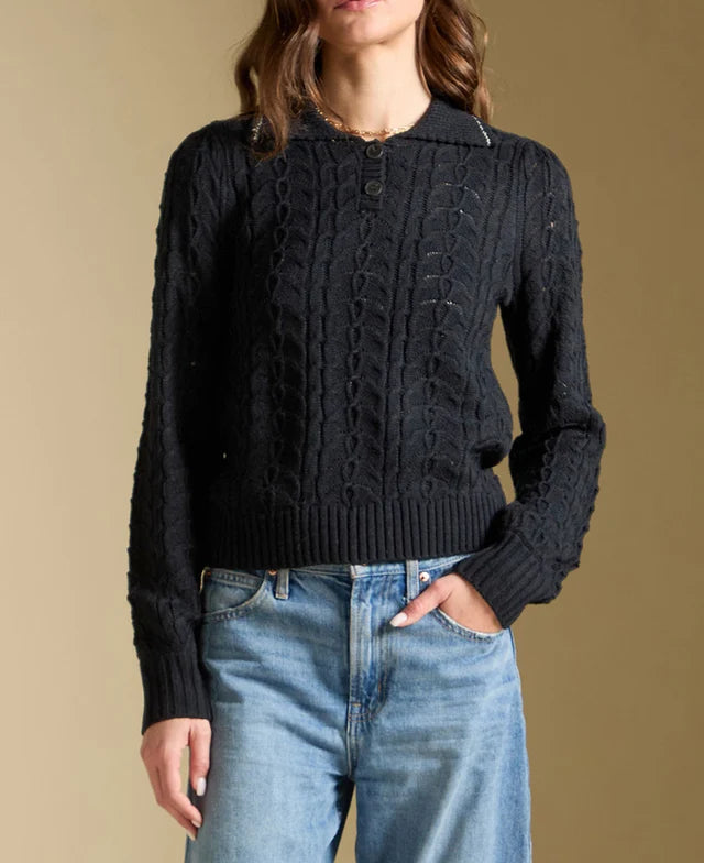 Woman wearing a black cable knit sweater and blue jeans against a beige background