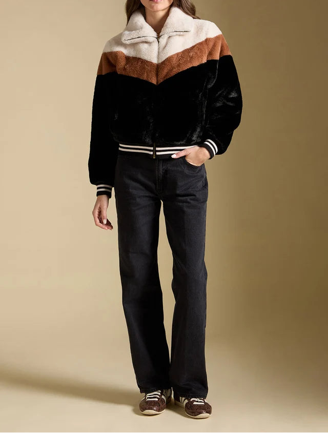 A model stands against a solid beige studio background wearing a color-blocked velvet jacket with a cream and brown chevron pattern, black sleeves, and striped ribbed trim, paired with dark jeans and brown sneakers.