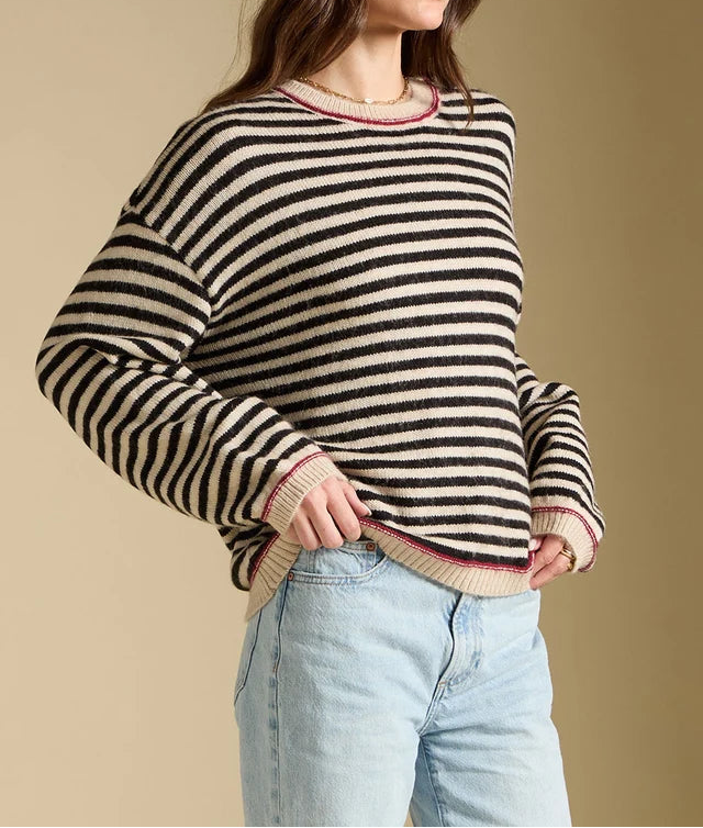 A woman wearing a black and beige horizontally striped knit sweater with pink trim, paired with light blue jeans, standing against a solid beige background.