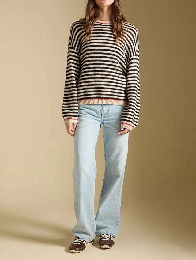 A woman modeling a black and white horizontally striped knit sweater, paired with light-wash wide-leg jeans and brown sneakers, against a solid beige studio background.