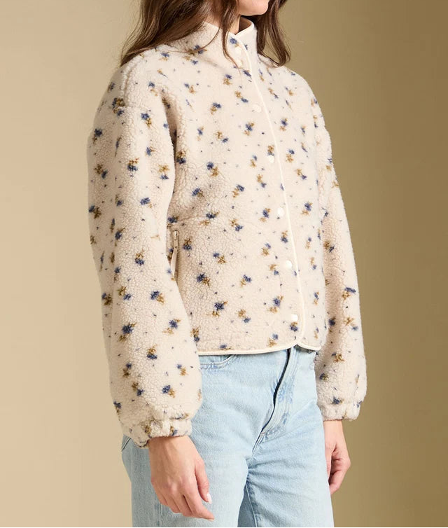 A woman wearing a cream-colored sherpa jacket with a small blue and yellow floral pattern, paired with light blue jeans, set against a solid beige background.