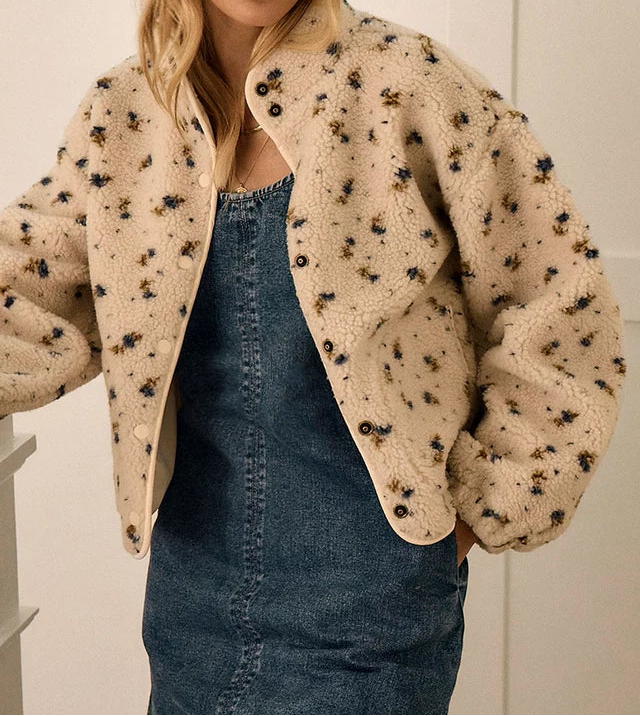 A woman wearing a beige sherpa-style cropped jacket with small black and brown floral embroidery, layered over a blue denim dress, set against a neutral indoor background.