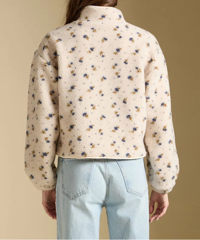 A woman viewed from the back wearing a cream-colored cropped fleece jacket with a small blue and yellow floral pattern, paired with light blue jeans, against a solid beige background.