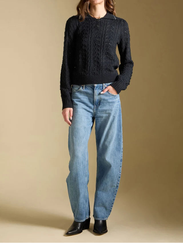 A woman stands against a warm beige studio background wearing a black cable-knit sweater, light-wash blue jeans, and black ankle boots.