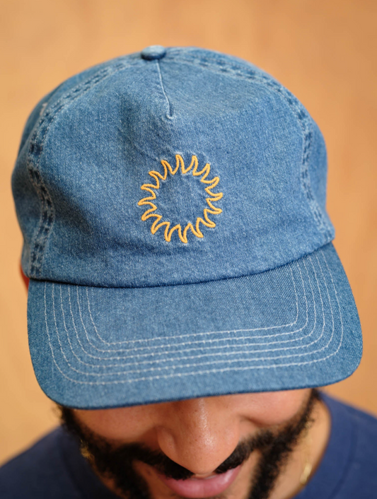 Blue denim cap with yellow embroidered design worn by a person against a beige background