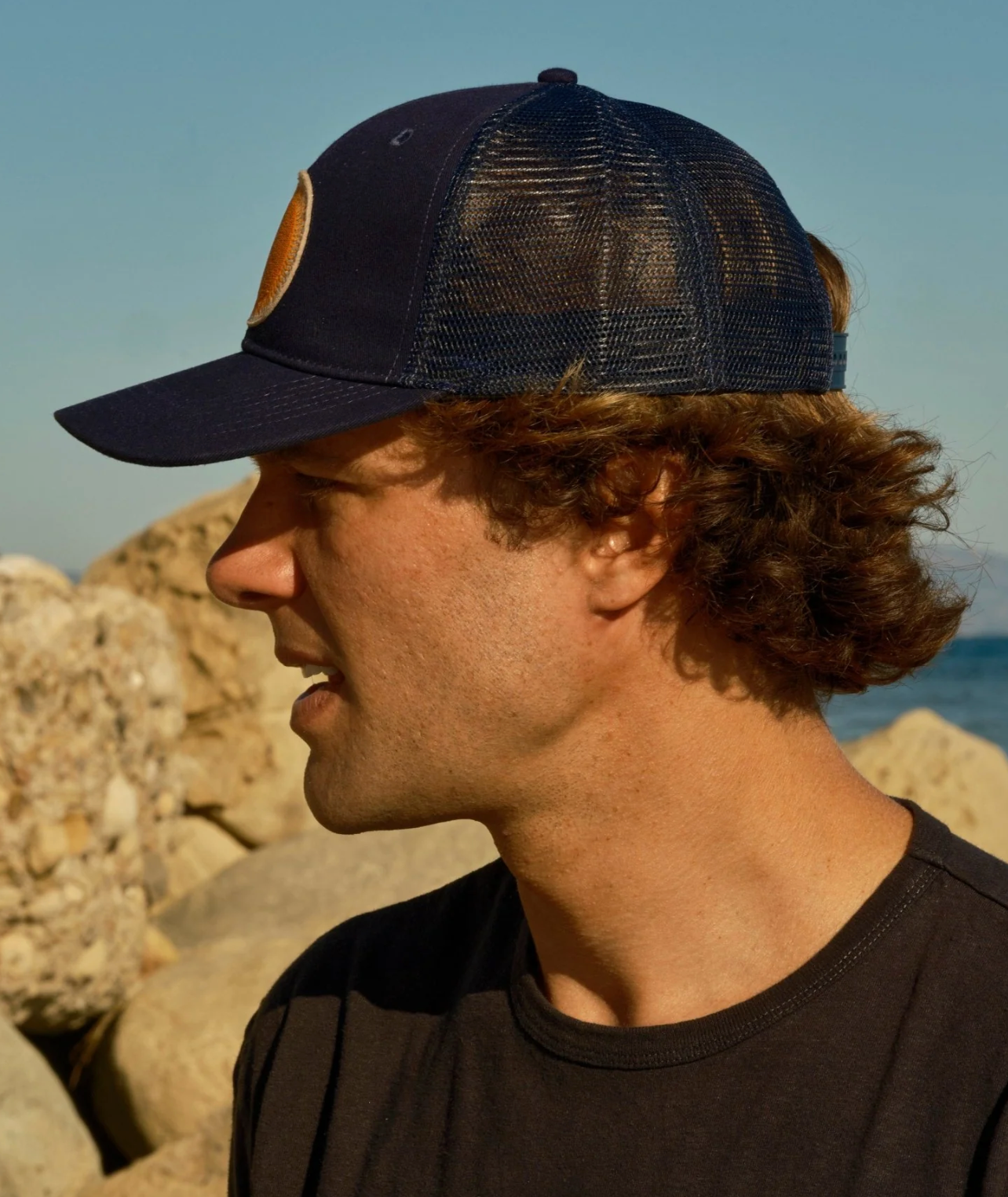 Man wearing a navy blue cap with mesh back, standing outdoors against a rocky background.