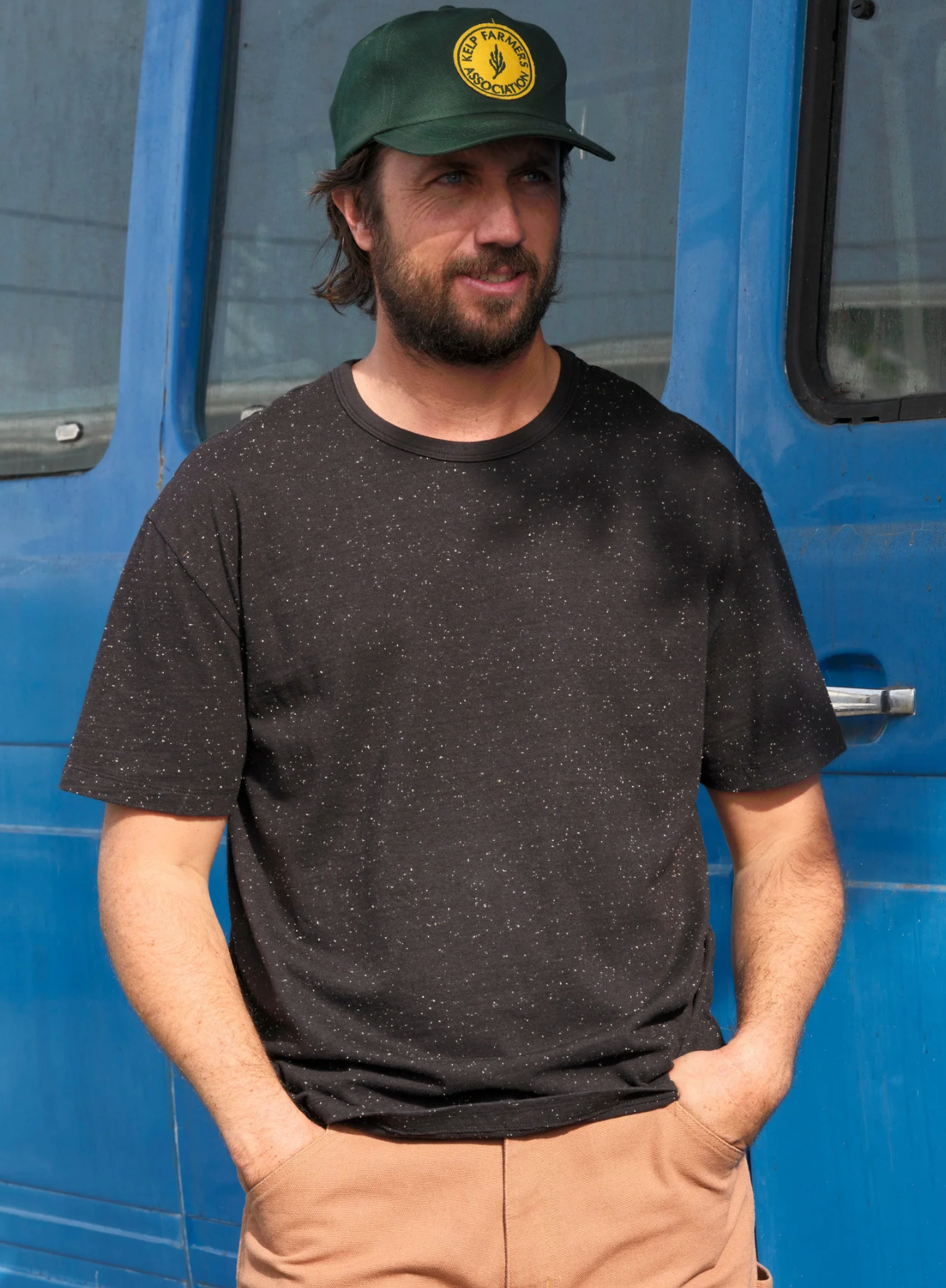 Man wearing a black t-shirt and green cap standing in front of a blue vehicle.
