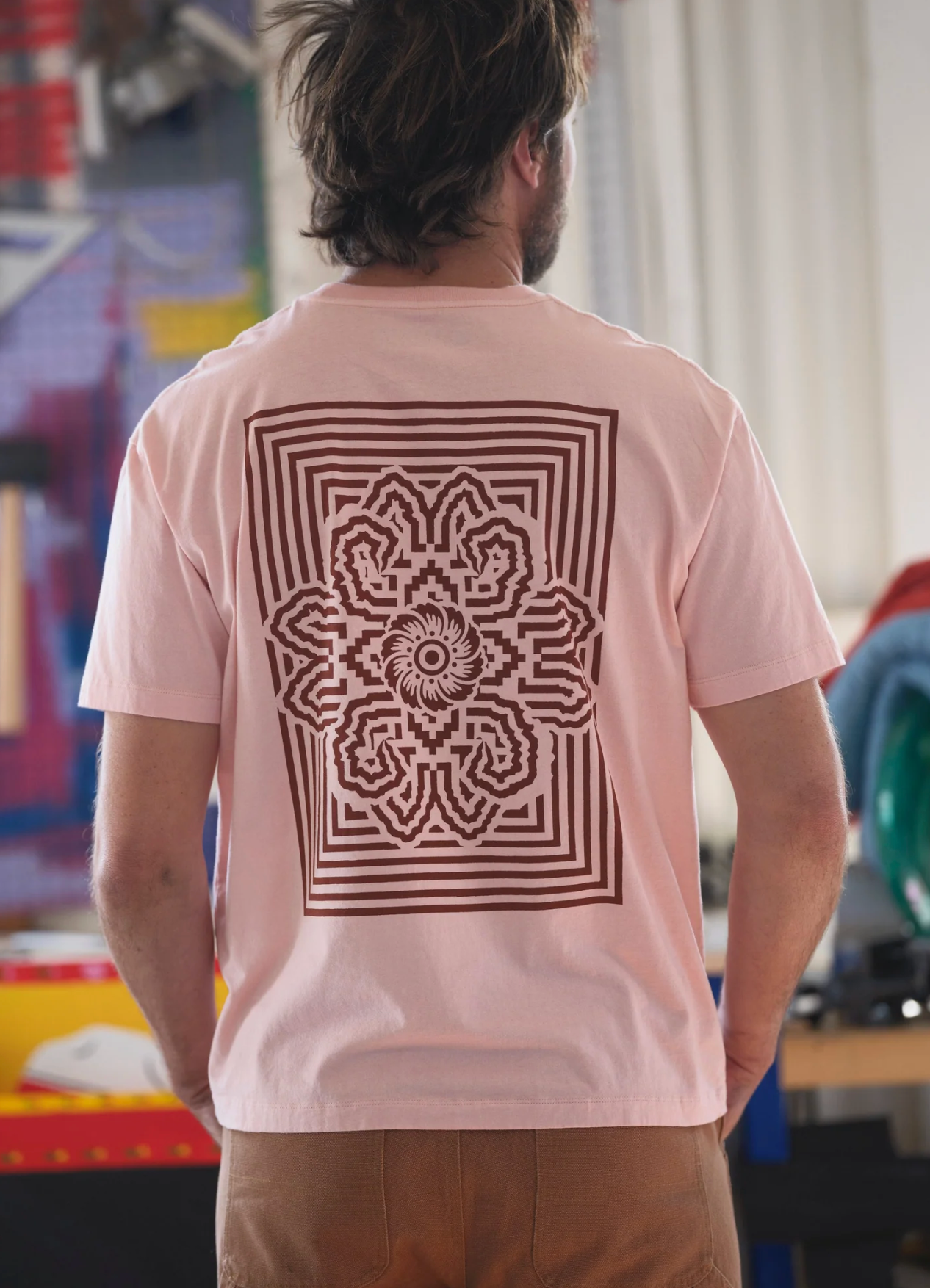 Person wearing a light pink t-shirt with a brown geometric design on the back, standing in an indoor setting.