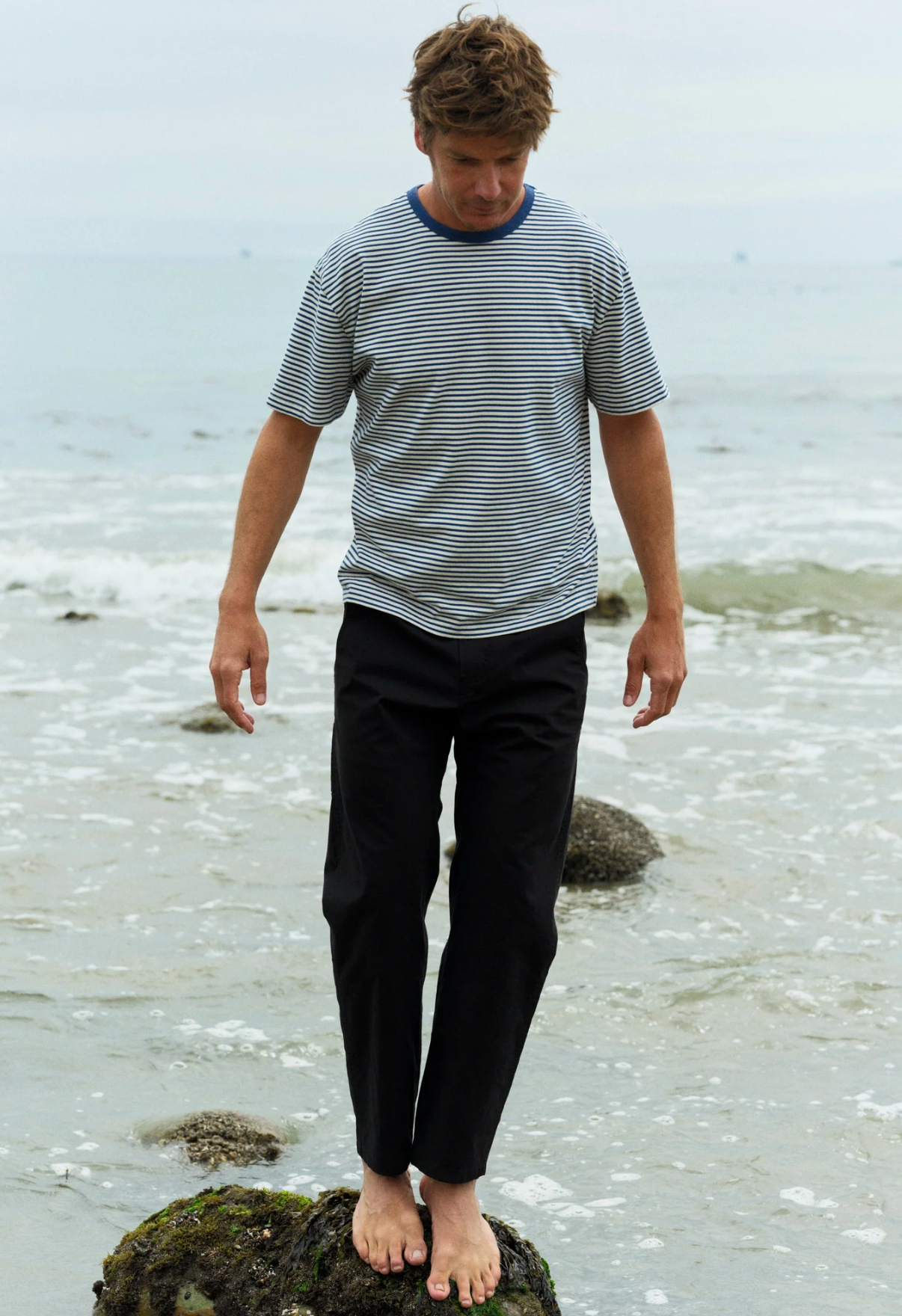 Man standing on a rock by the water's edge wearing a striped shirt and dark pants.
