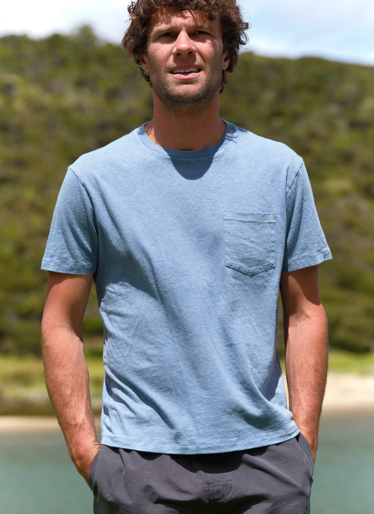 Man wearing a light blue t-shirt with a pocket, standing outdoors with greenery in the background.