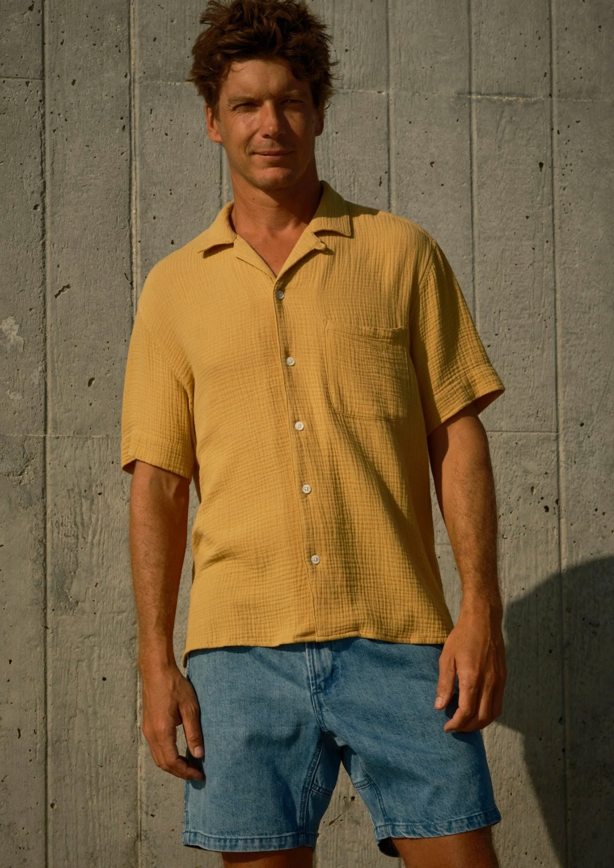 Man wearing a yellow shirt and blue shorts standing against a concrete wall.