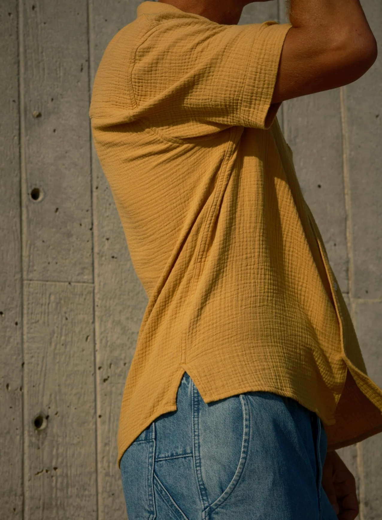 Person wearing a yellow textured shirt and blue jeans against a concrete wall.