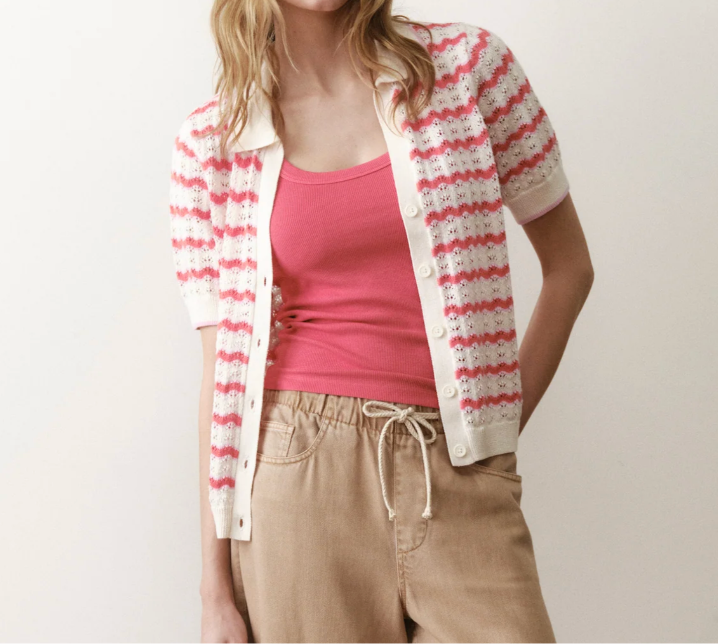 Woman wearing a pink top and beige pants with a white and red striped cardigan on a plain background