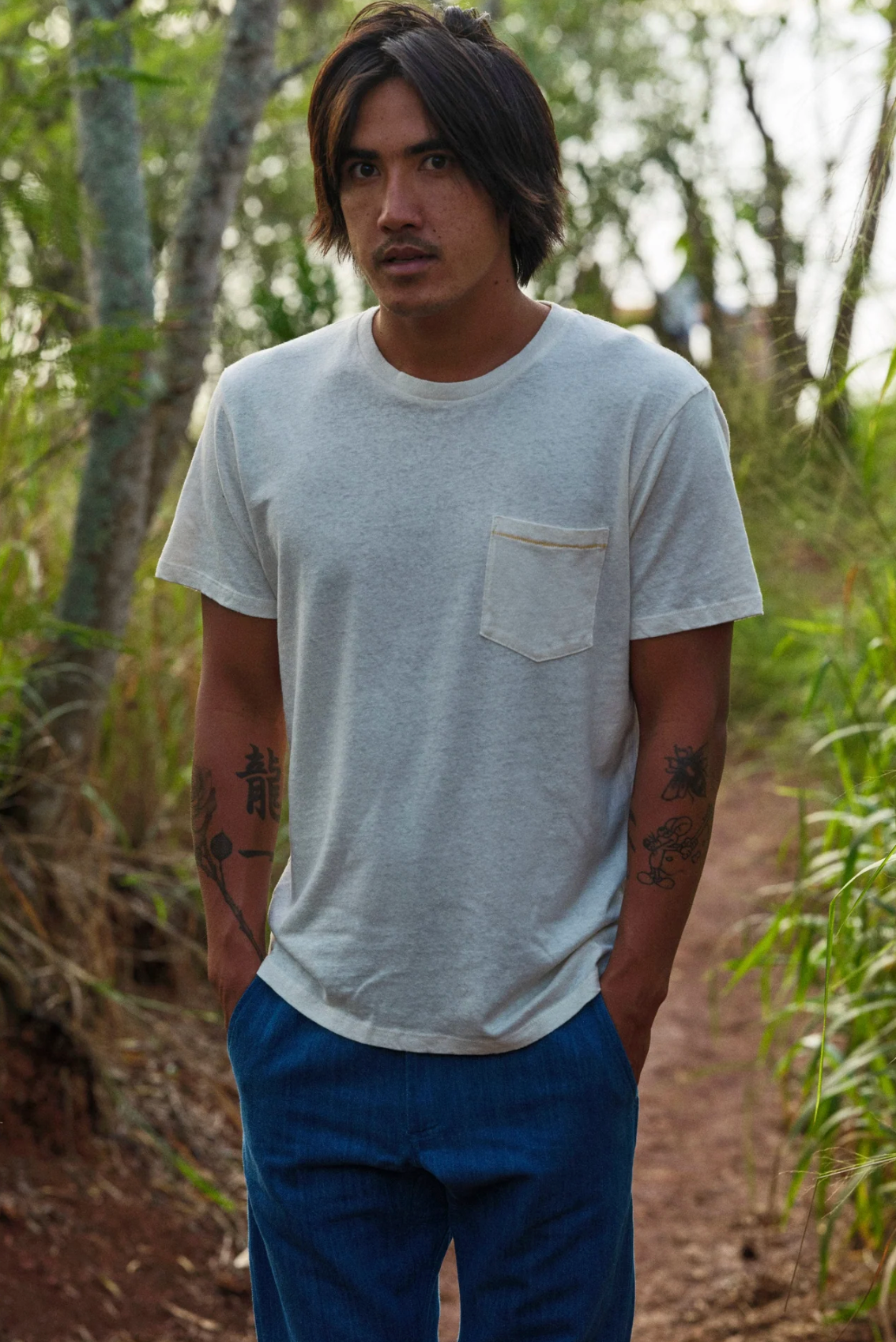 Man wearing a light gray t-shirt with a pocket and blue pants standing in a forest.