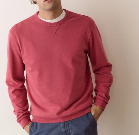 A man wearing a dusty rose crewneck sweatshirt over a white t-shirt and blue pants, standing against a plain beige wall.