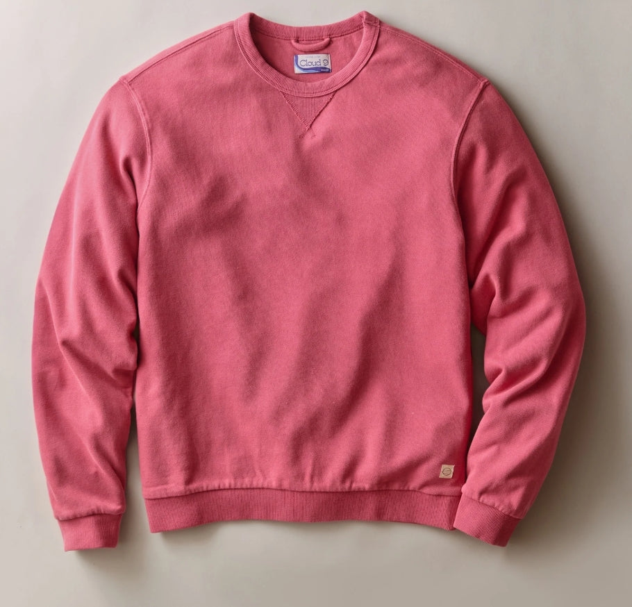 A pink crewneck sweatshirt laid flat against a neutral light gray background. The sweatshirt has a classic crew neck, long sleeves, ribbed cuffs and hem, and a small tan brand patch near the bottom right hem.