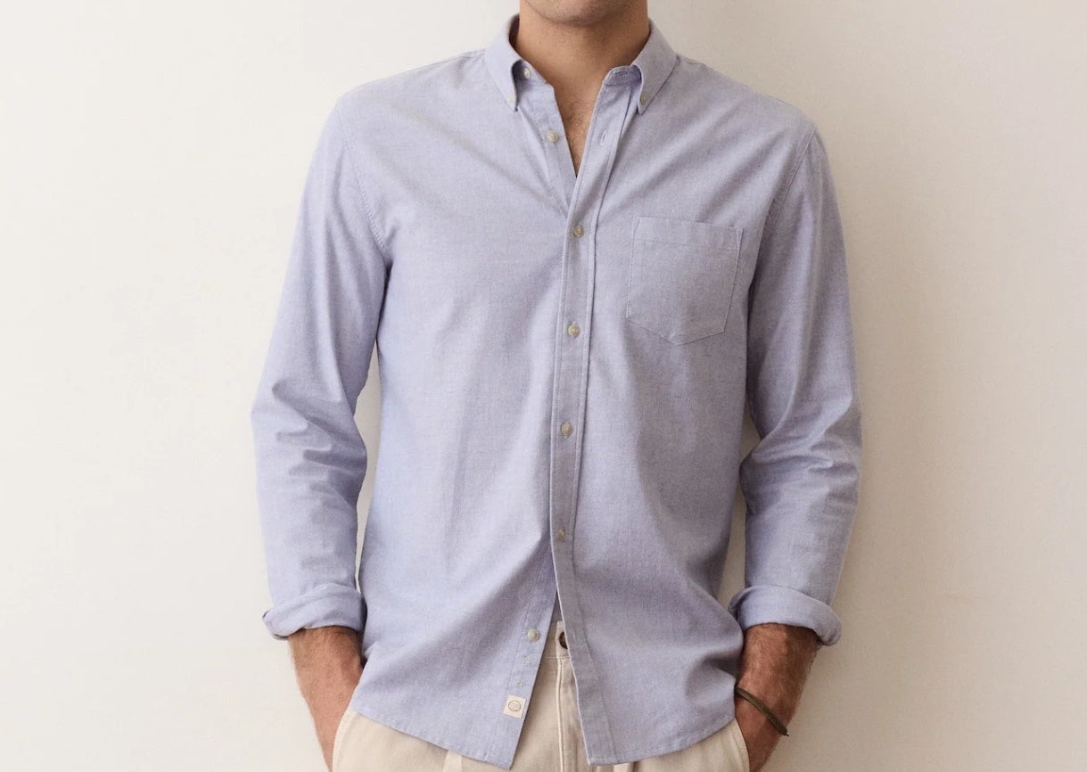 A man wearing a light blue long-sleeve button-down shirt with rolled-up sleeves and beige pants, standing against a plain off-white wall.