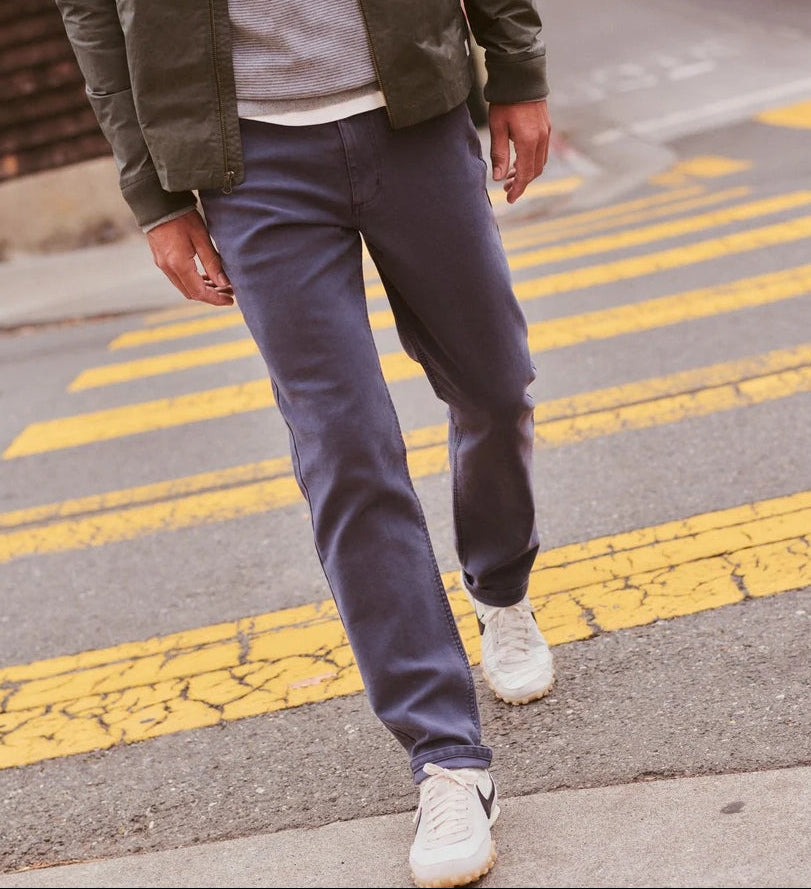 A person outdoors wearing slim straight-fit charcoal gray pants, styled with a dark olive jacket, gray shirt, and white sneakers. The image includes a vertical panel showing color and fit options for the pants.