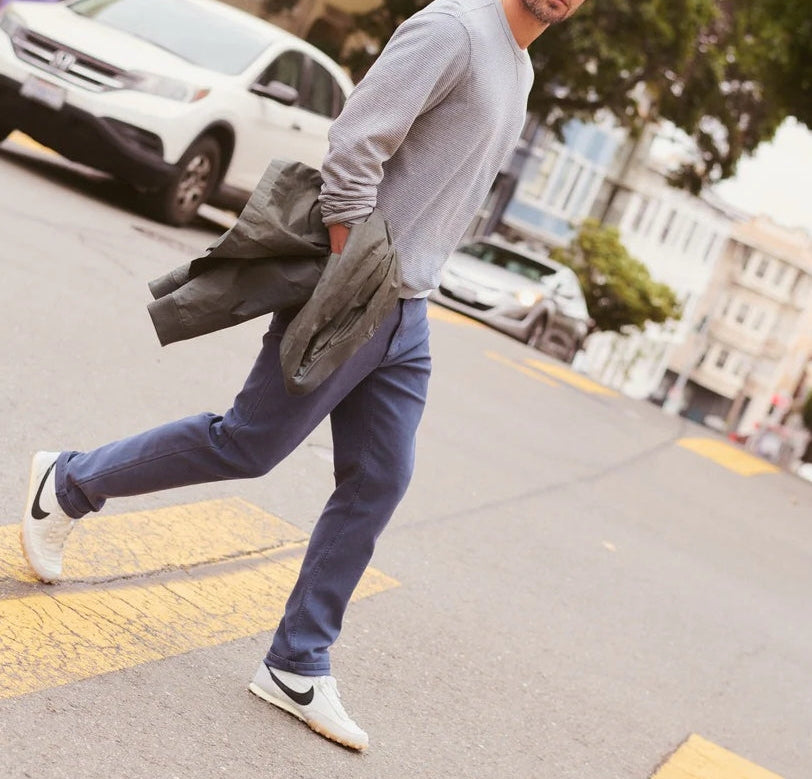 A man walking on a city street wearing a Flex Terry straight-leg pant in navy blue, a light gray long-sleeve shirt, and white sneakers, holding a dark jacket. The image is a lifestyle product shot with text overlays at the top and bottom.