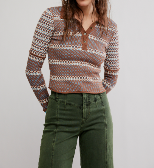 A woman wearing a brown, beige, and white patterned knit long-sleeve henley top with olive green pants, photographed against a plain light gray studio background.