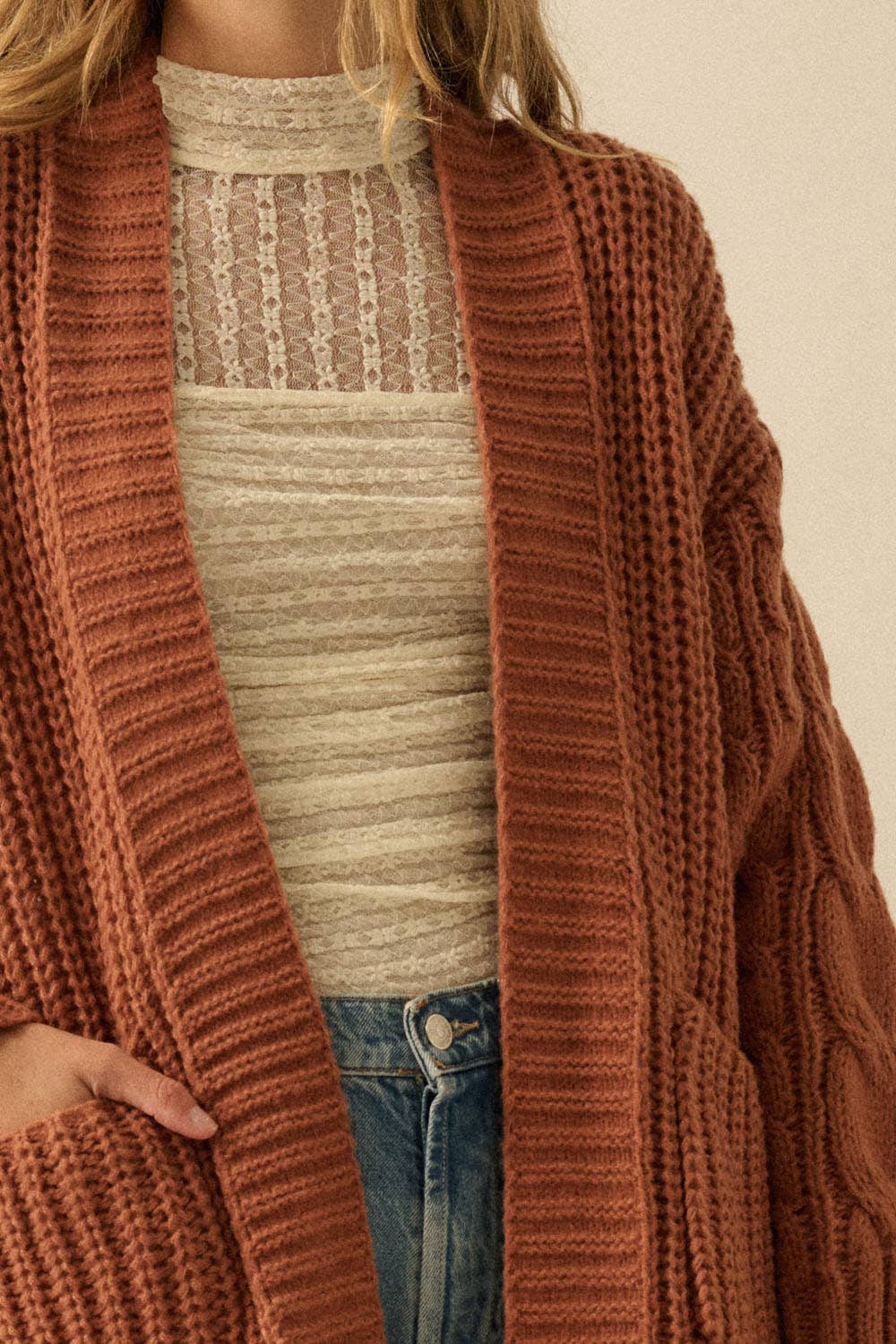 A close-up of a person wearing a chunky open-front cardigan in a warm rust or terracotta color over a cream ribbed top and blue jeans.