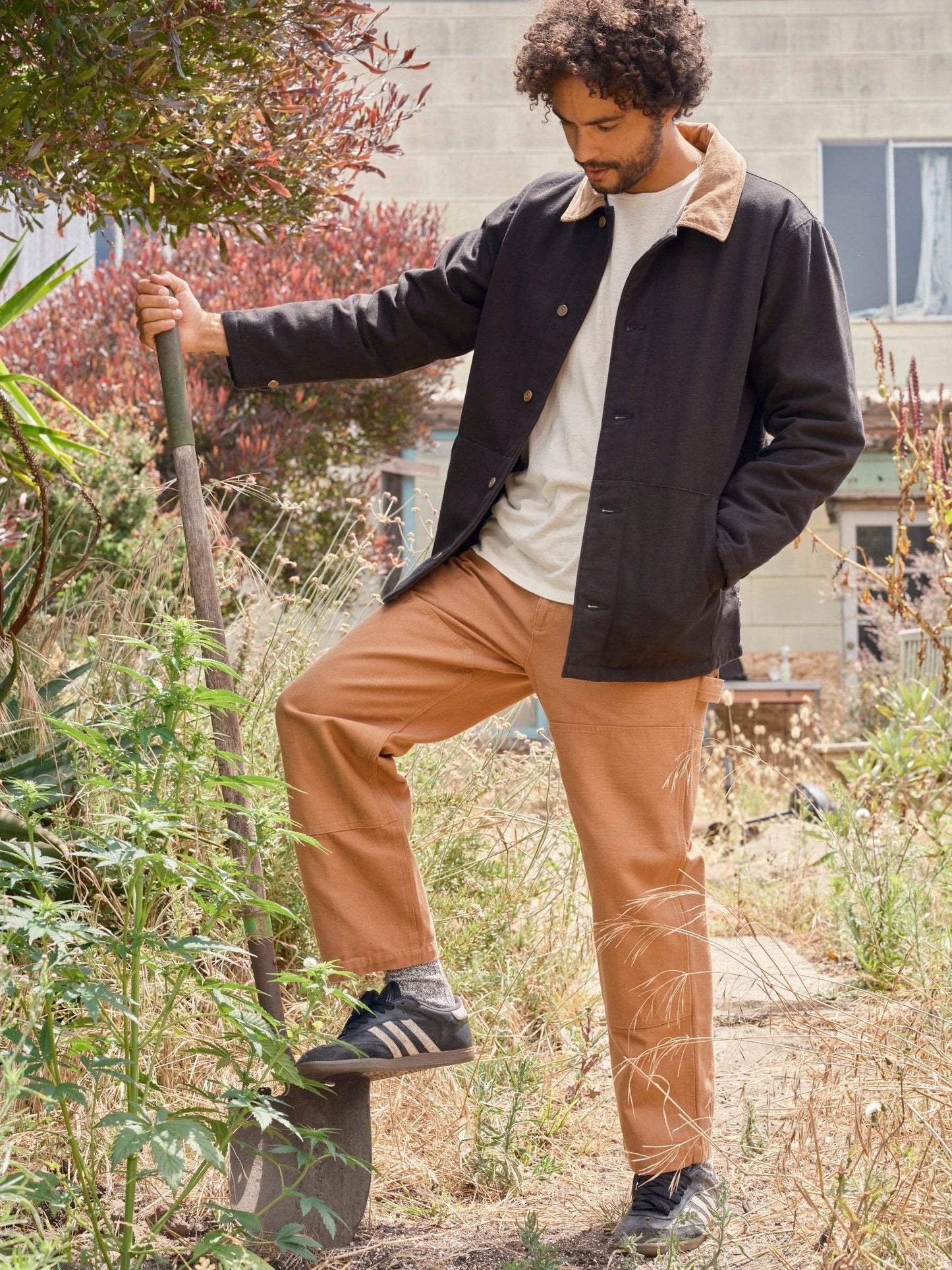 Man standing in a garden holding a shovel, wearing a black jacket and brown pants.