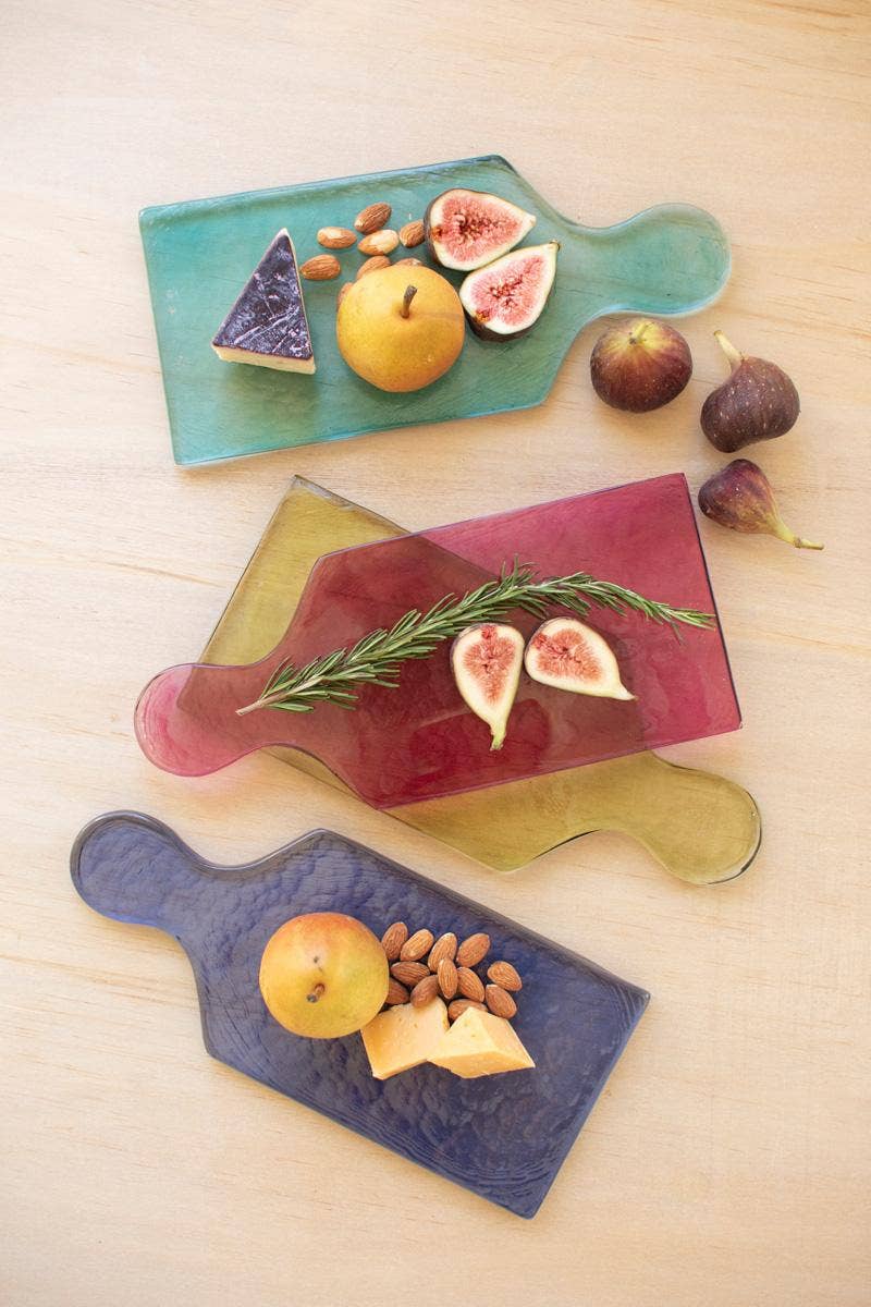 Three hammered glass serving boards in teal, translucent olive green, and dark navy blue, styled with figs, almonds, and cheese on a light wood background.
