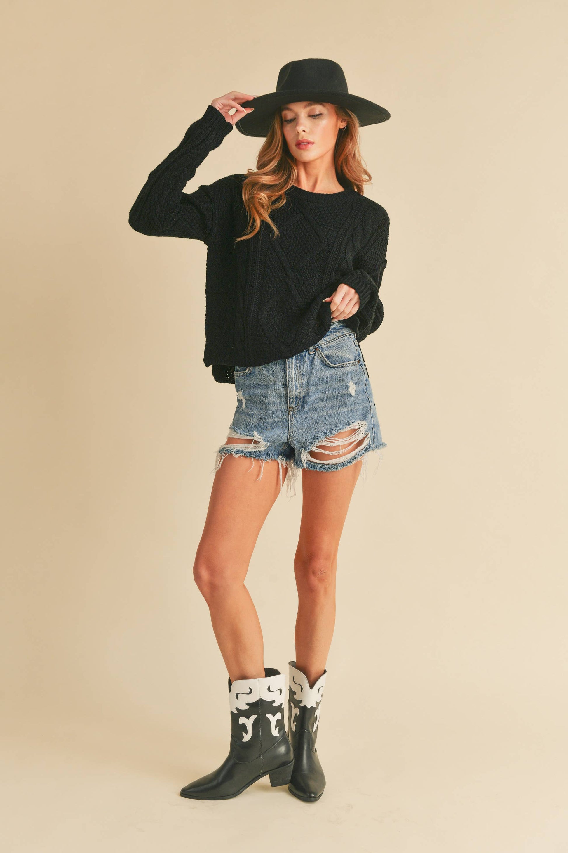 Woman wearing a black sweater, denim shorts, and cowboy boots with a black hat on a beige background