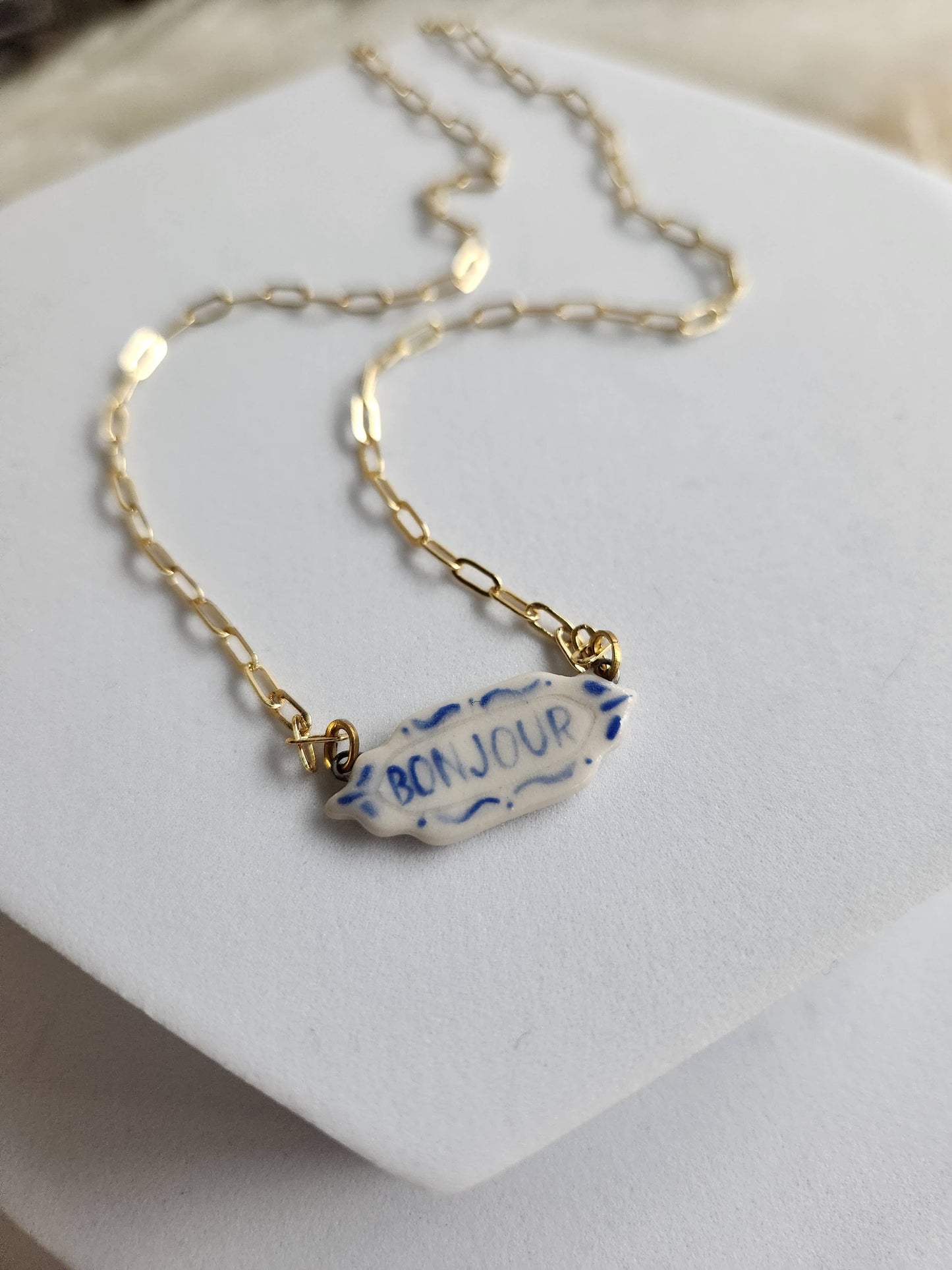 Gold necklace with a 'Bonjour' tag on a white stand