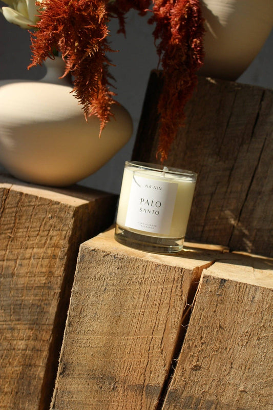 A candle in a clear glass jar with a cream-colored wax and a white label reading 'Palo Santo' and 'BURN', styled on rustic wooden blocks with dried orange-red foliage and neutral ceramic vases in the background.