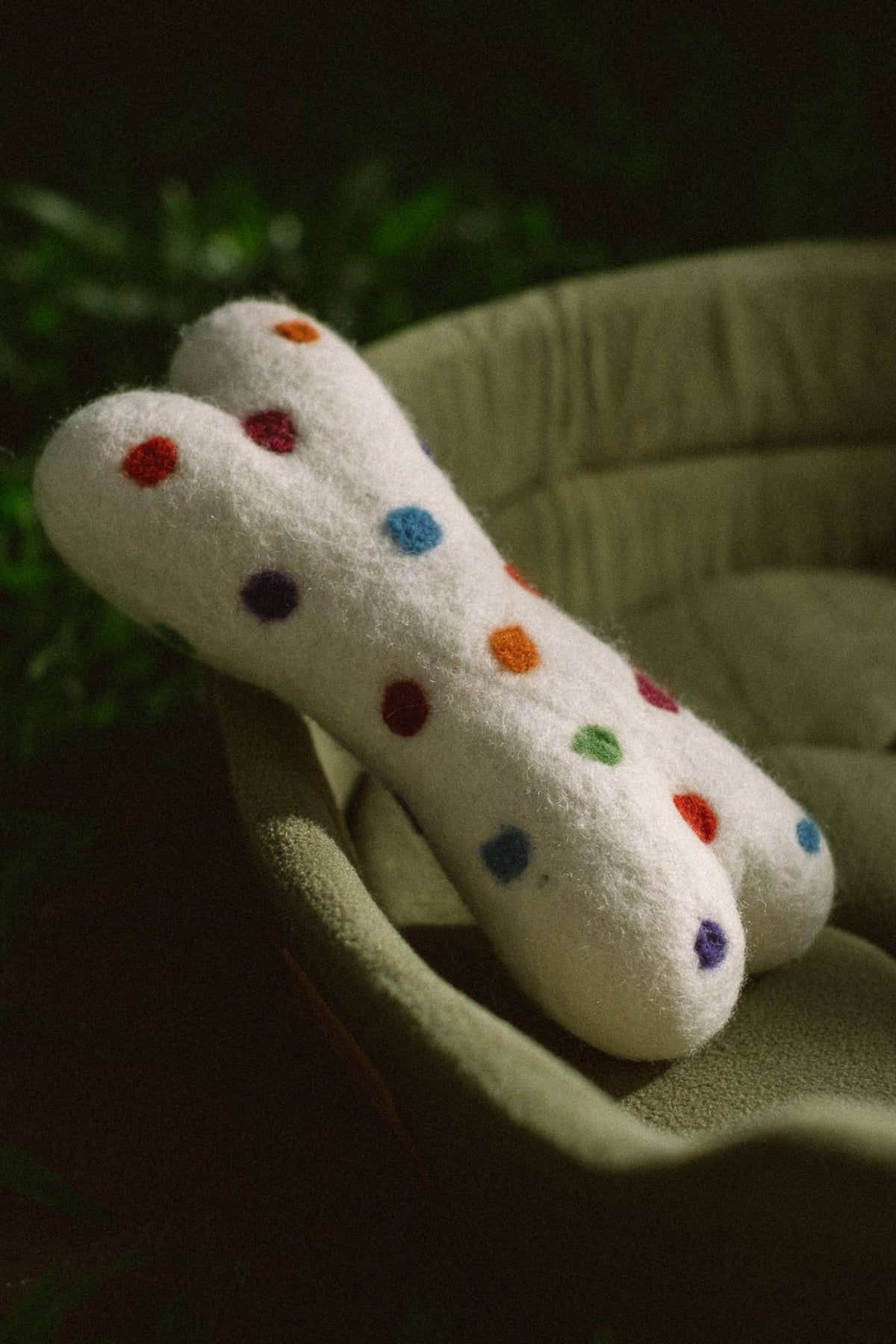 A white felt pet toy shaped like a bone, decorated with colorful embroidered polka dots, resting on a green cushioned surface.