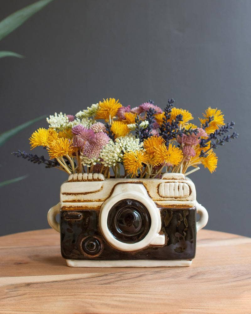 Ceramic camera-shaped vase with flowers on a wooden surface against a dark background
