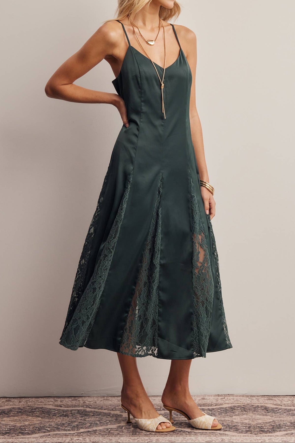 A woman models a dark green midi slip dress with thin straps, a lace overlay, and a long gold pendant necklace, standing against a neutral beige wall.