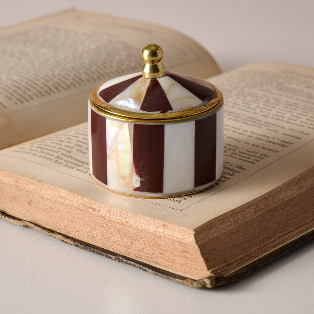 A small decorative lidded box with a striped pattern of deep burgundy and white mother-of-pearl, accented with gold trim and a gold finial, resting on an open vintage book.