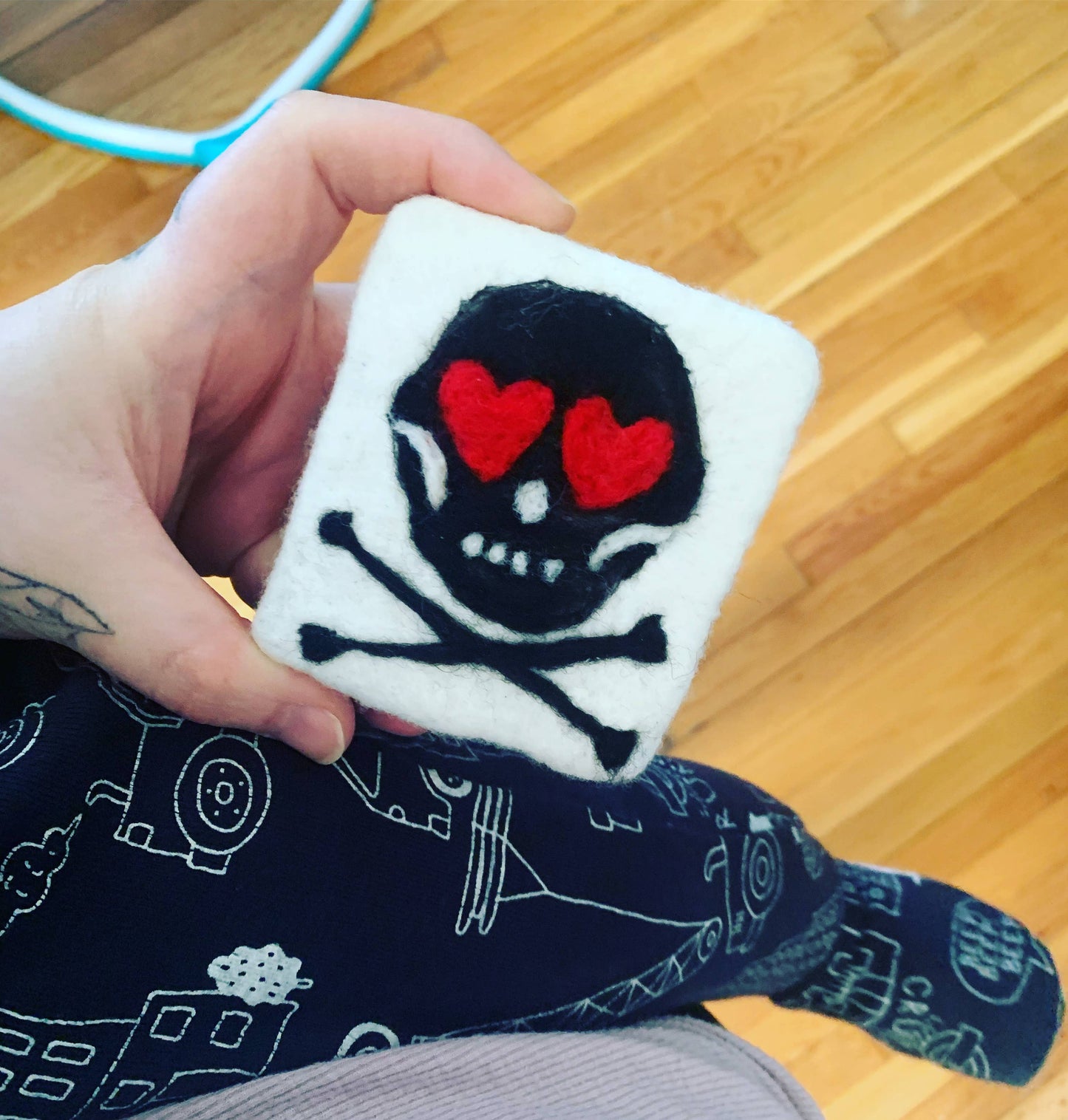 Hand holding a small square plush toy with a skull and crossbones design featuring red hearts on a wooden floor.
