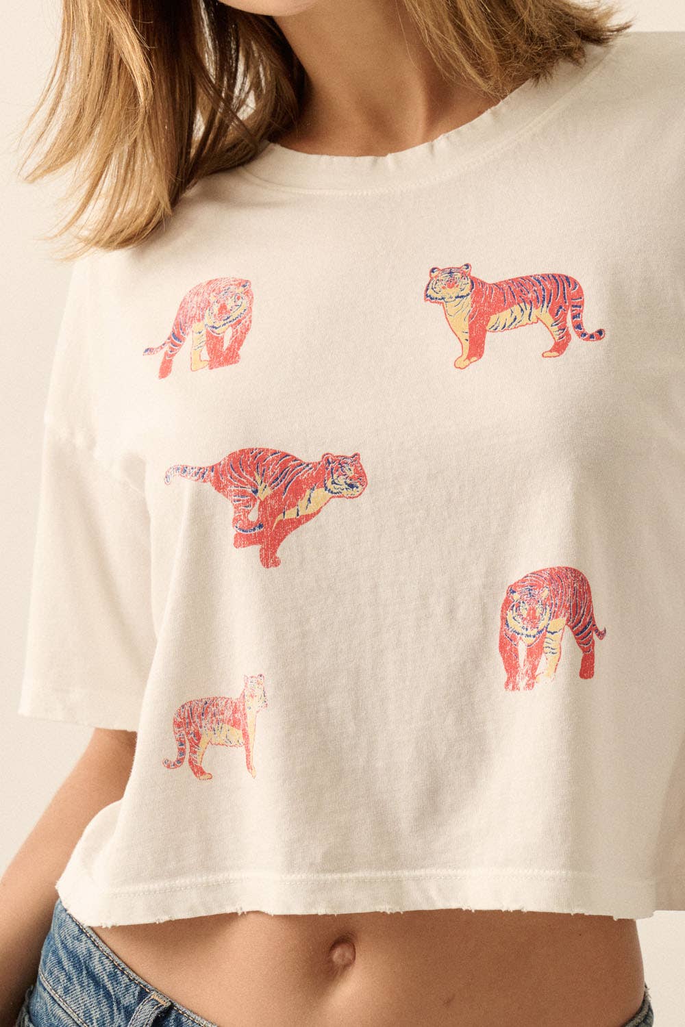 A cropped off-white t-shirt featuring a pattern of stylized tigers in red, orange, and yellow, worn by a model.