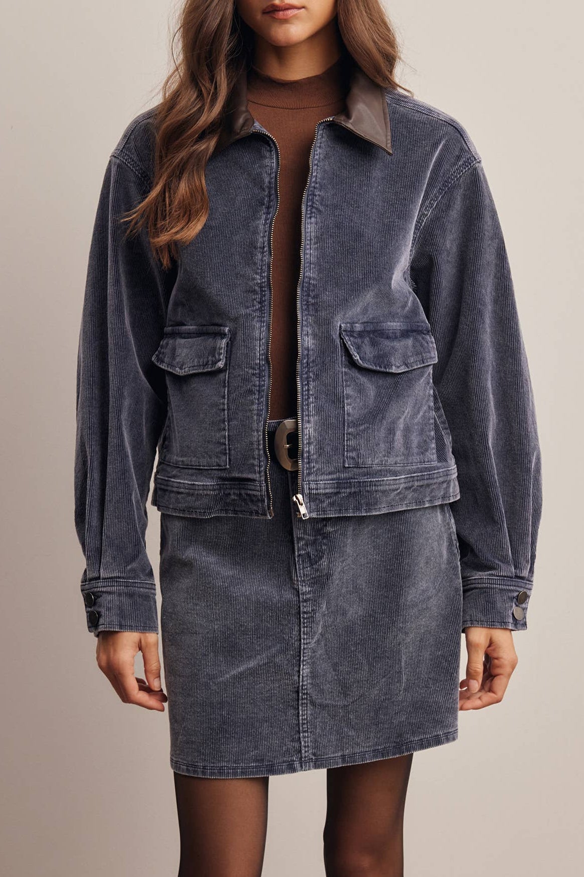 A model wearing a dark blue corduroy jacket and matching corduroy mini skirt with brown turtleneck and brown boots, standing against a solid beige background.