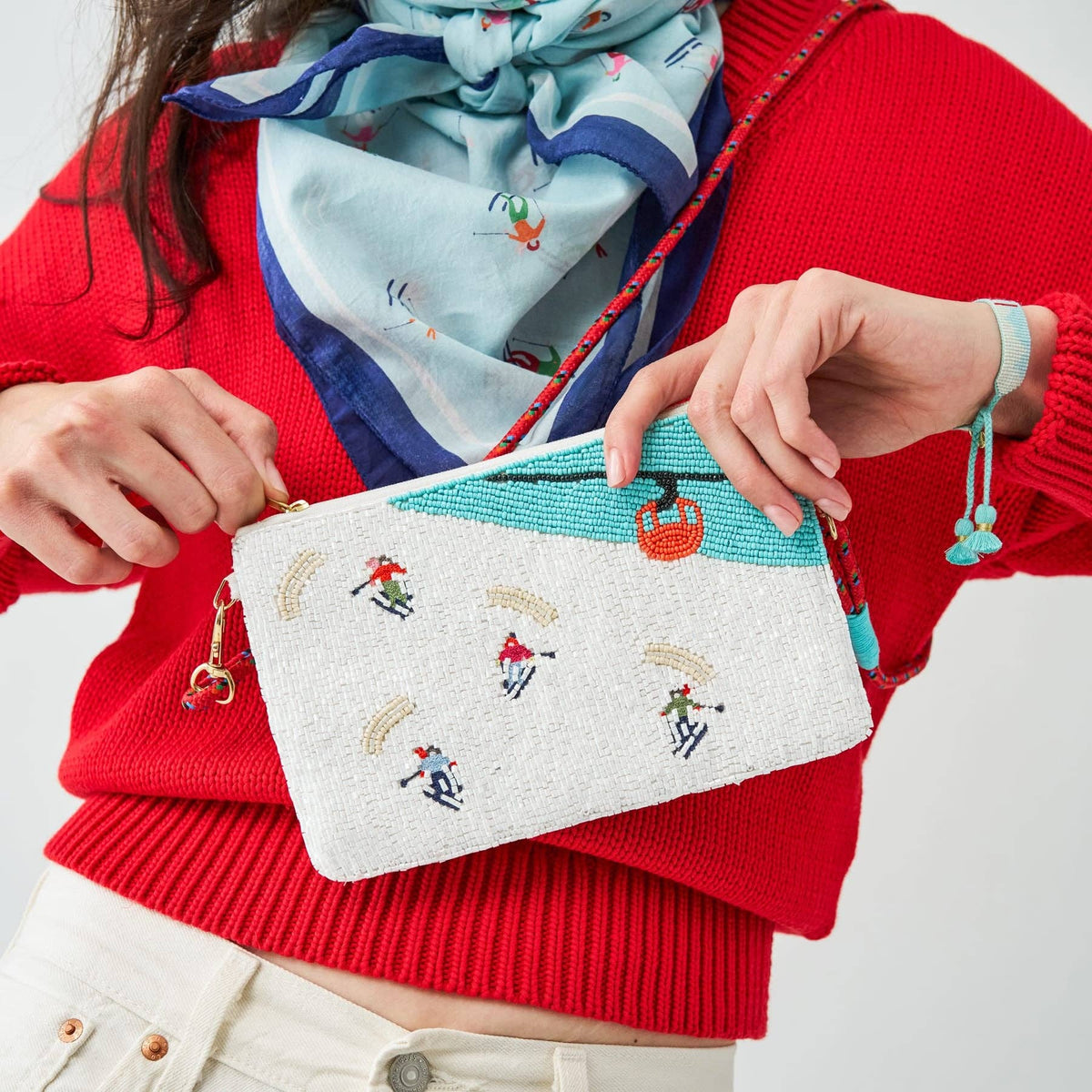 A person wearing a bright red sweater and a light blue patterned scarf holds a white embroidered crossbody pouch with colorful ski-themed needlepoint details. The pouch has a turquoise stripe, a small red gondola, and embroidered skiers.
