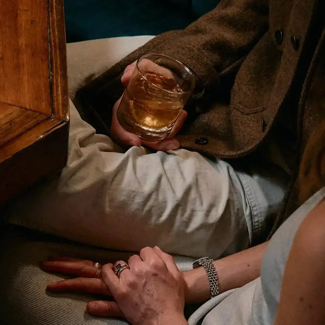 Person holding a glass of amber liquid with another person's hand on a wooden surface.