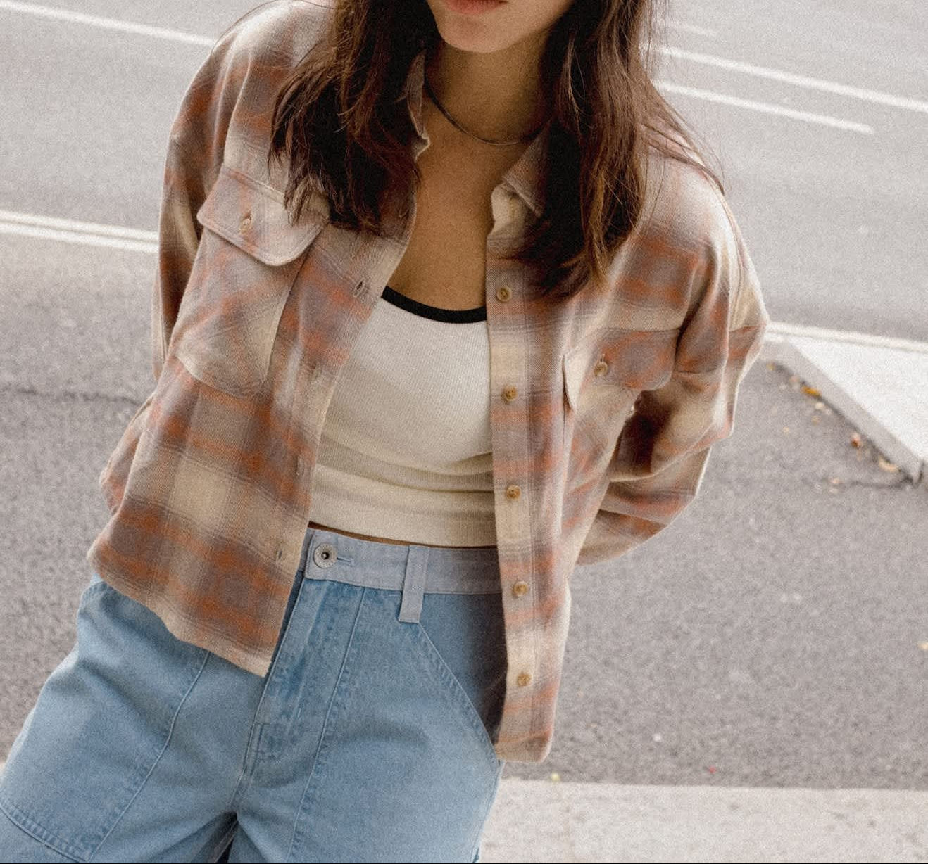 A woman wearing a cream cowboy hat, a brown and beige plaid flannel shirt over a white tank top, and light blue jeans, standing on a city street.