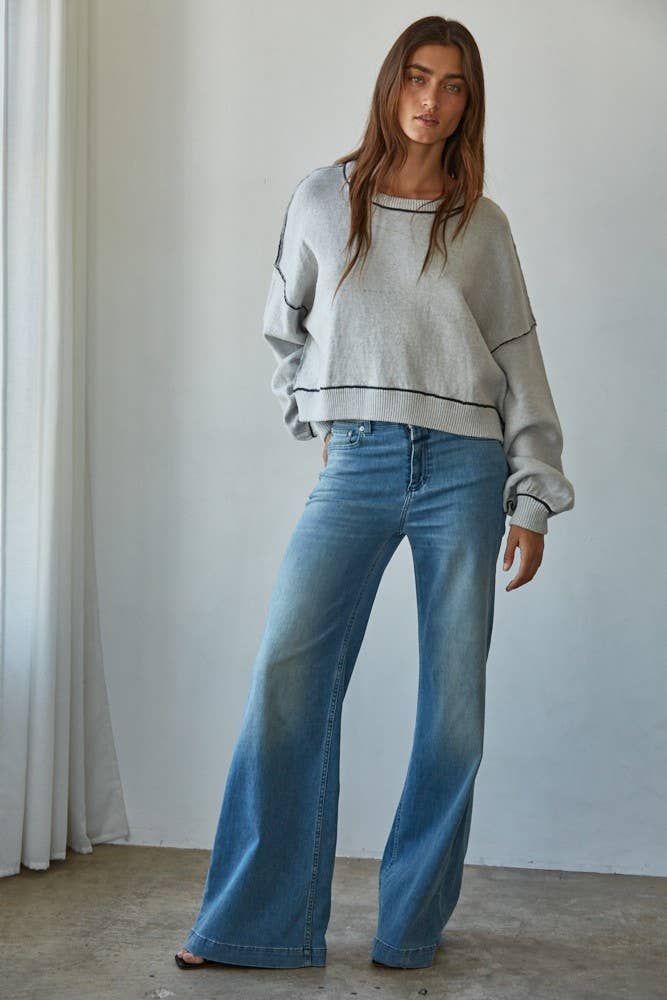 Woman wearing a light gray sweater and blue jeans standing against a white wall.