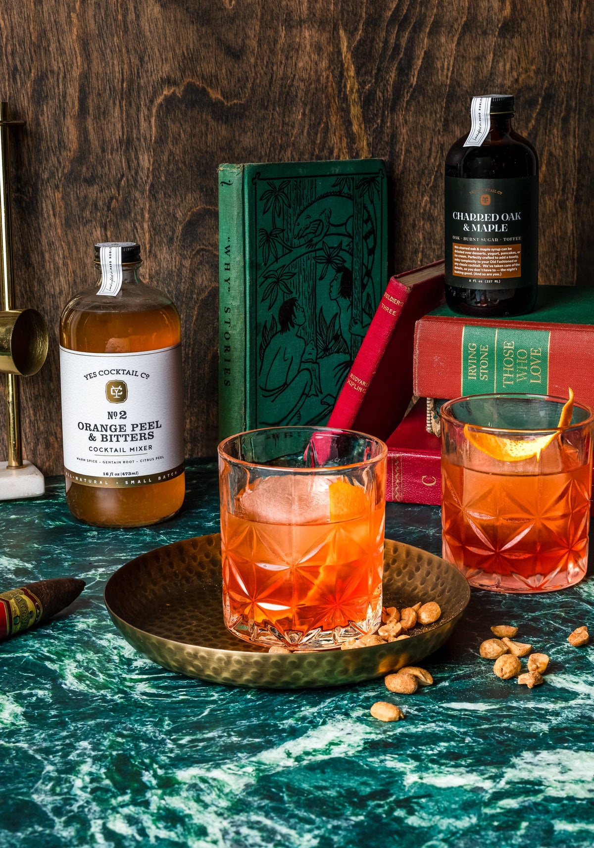 Two glasses of orange cocktail with ice on a wooden tray, surrounded by bottles and books on a dark surface.