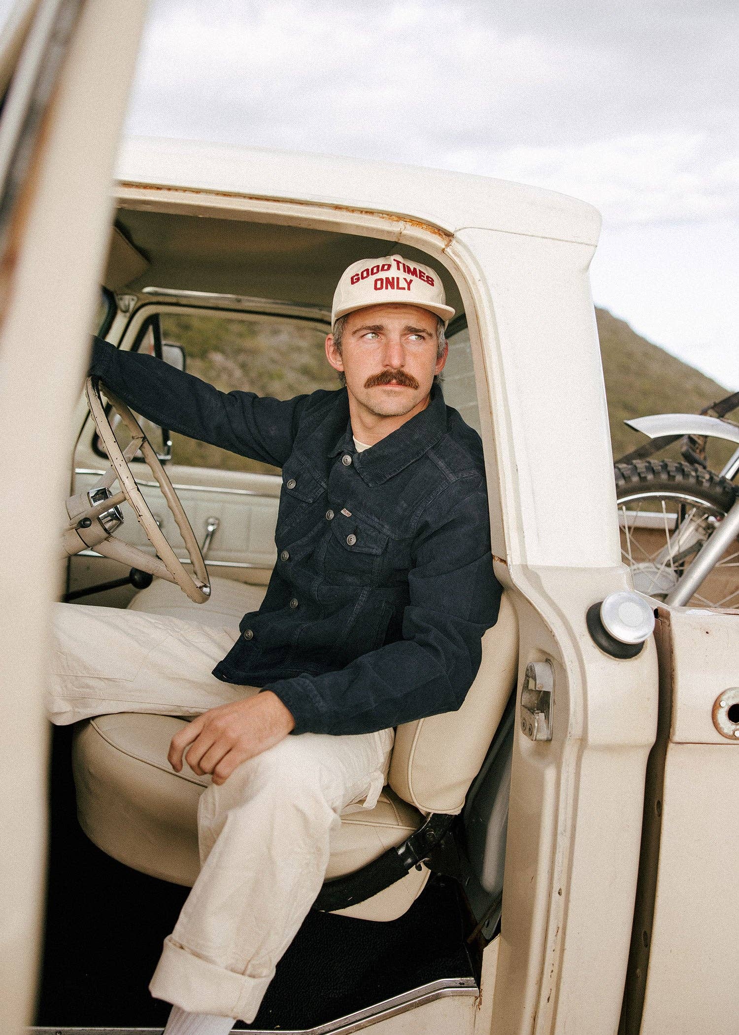 Man sitting in a vintage car wearing a cap and denim jacket.