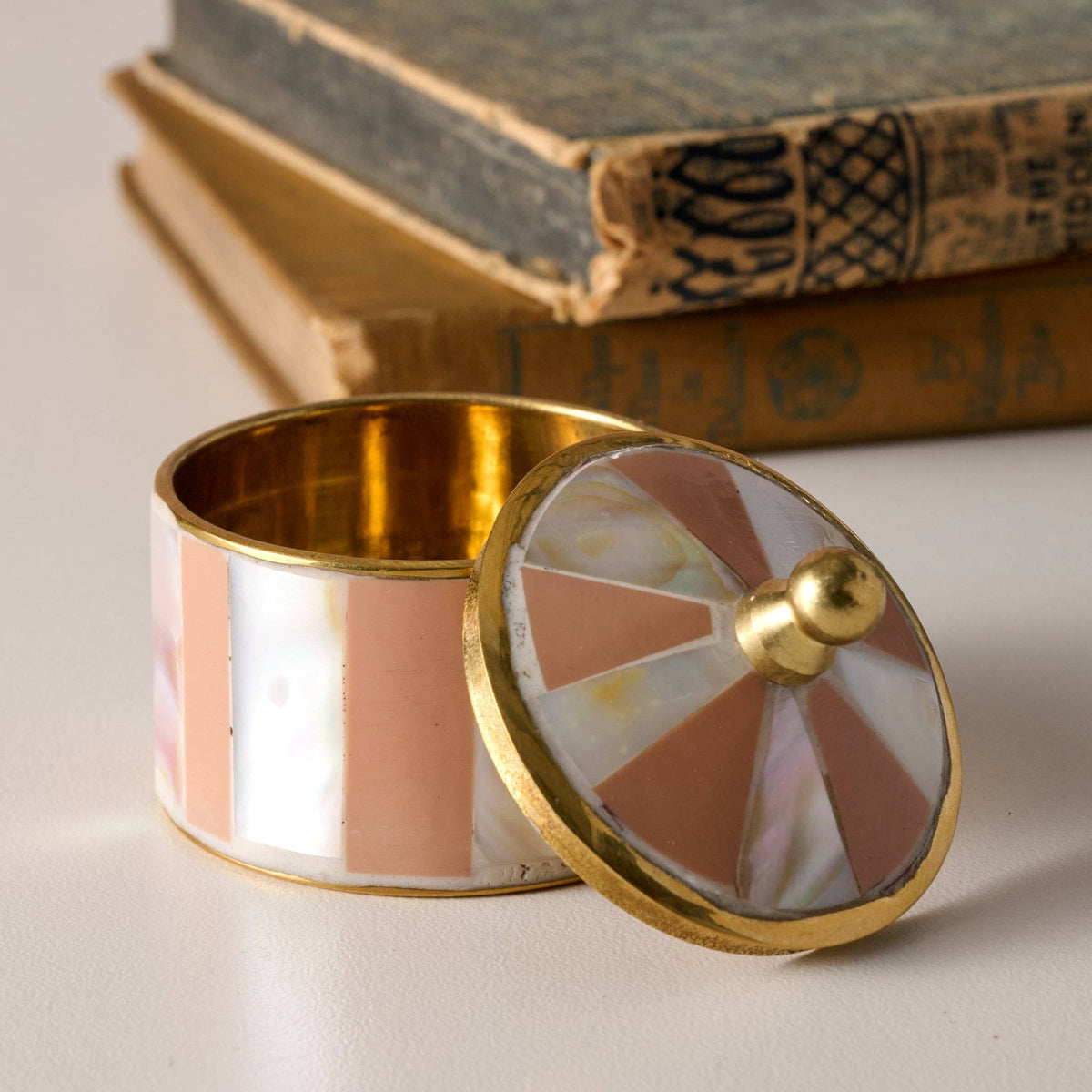 A small cylindrical jewelry box with a matching lid, featuring a gold-tone brass frame and inlay of pearlescent white and dusty pink mother-of-pearl segments. The lid is propped open next to the box, which is set against a soft-focus background of stacked vintage books.