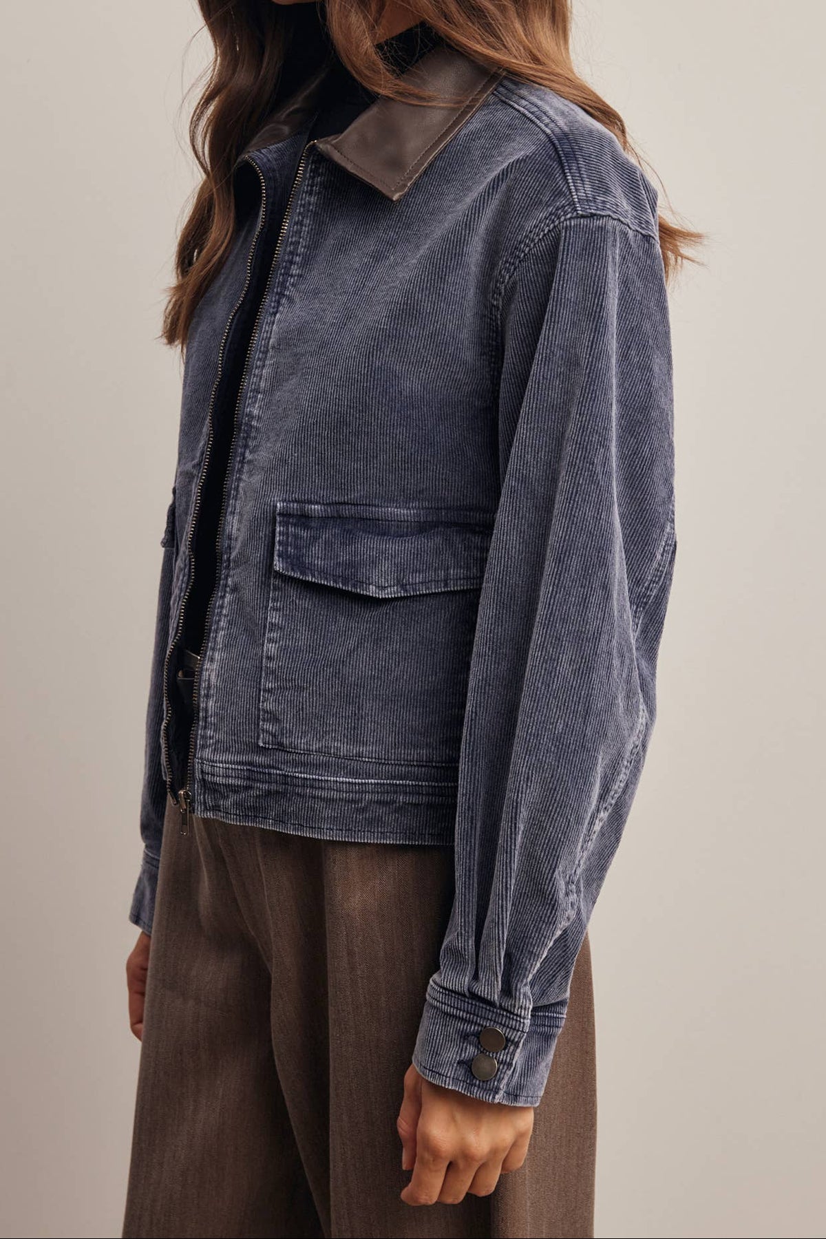 A woman wearing a dark blue-gray corduroy jacket with a brown collar, paired with brown trousers, photographed against a neutral beige background.