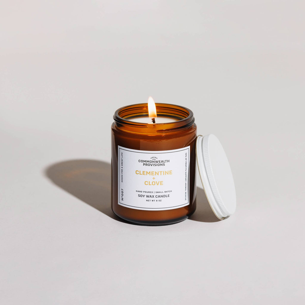 An amber glass jar candle with a white label reading 'CLEMENTINE + CLOVE' and a lit flame, shown on a light gray studio background with the white lid resting beside the jar.