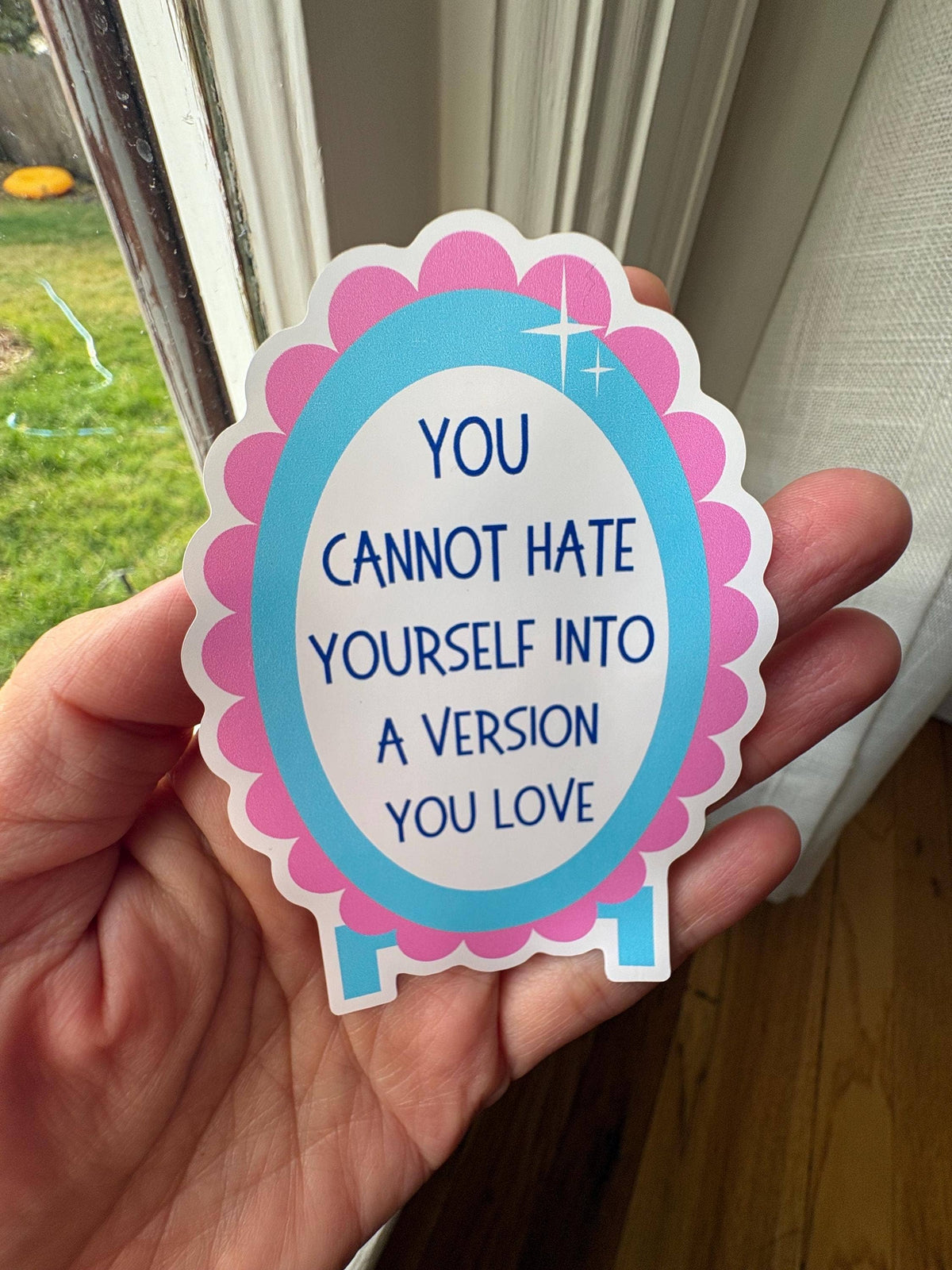 A hand holds a die-cut sticker with the text 'YOU CANNOT HATE YOURSELF INTO A VERSION YOU LOVE' in blue lettering. The sticker has a white center, a light blue oval border, and a scalloped pink outer border.