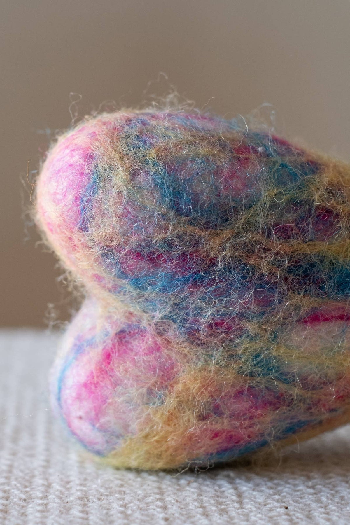 A close-up of a fuzzy, heart-shaped felted wool object in marled pastel colors including pink, blue, yellow, and cream, resting on a textured light fabric surface against a soft beige background.