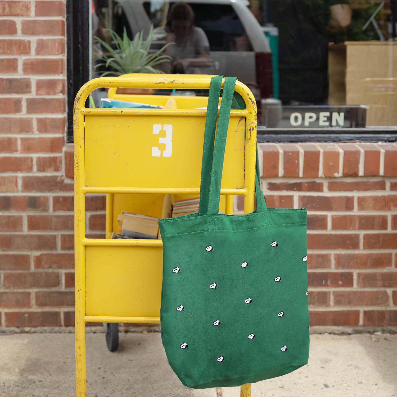 Green tote bag with white embellishments hanging on a yellow post box against a brick wall.