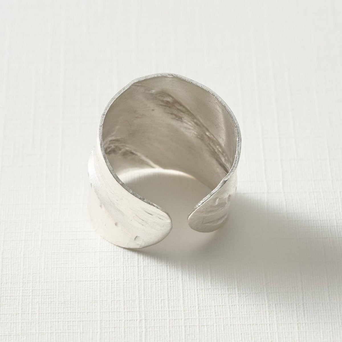 A hammered silver cuff ring with a wide, open bangle design, photographed on a textured white surface.